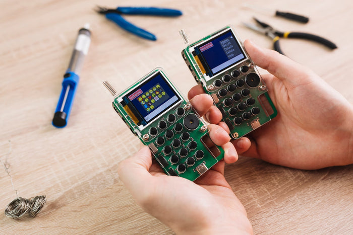 Two green handheld DIY walkie-talkies with screens and antennas held over a workbench with electronics tools.