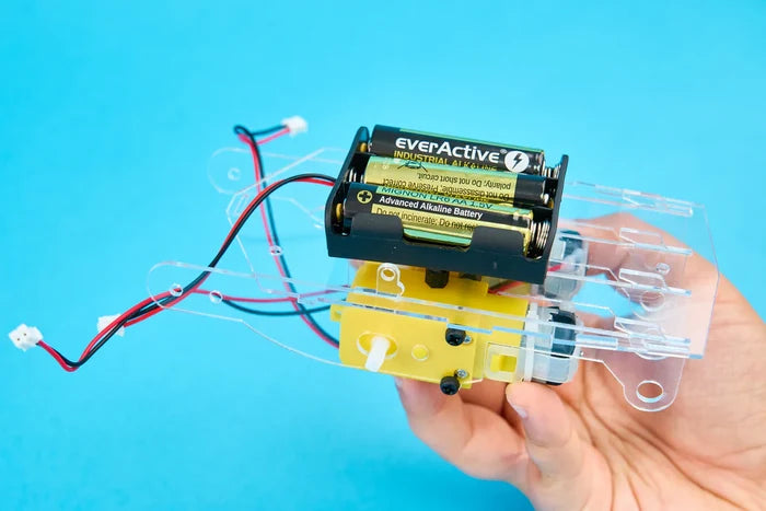 Hand holding a clear acrylic robot base with yellow gear motors, AA batteries, and wiring against a blue background.