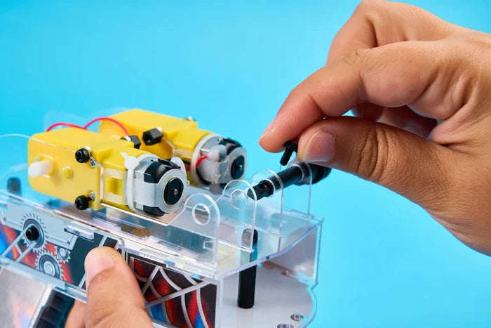 A person's hands assemble a yellow and clear plastic robot toy against a blue background, holding a small black screw.