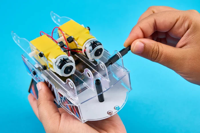 Hands assembling a clear plastic robot with yellow motors and red wires using a screwdriver on a blue background.