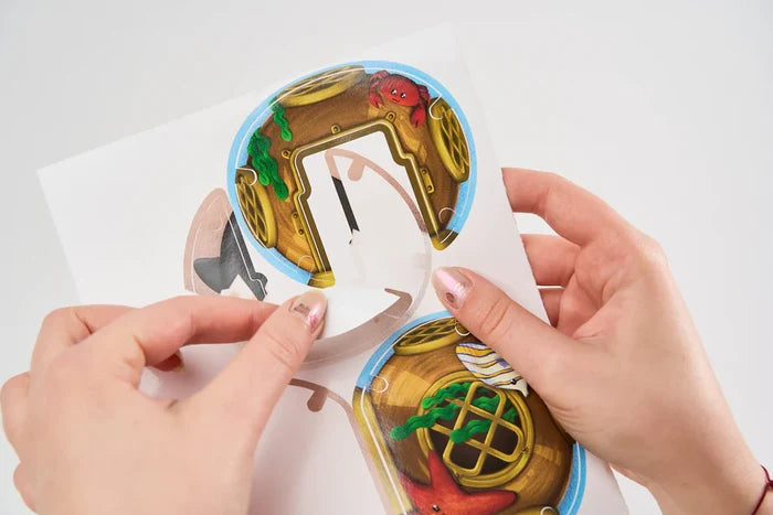 Hands peeling a circular sticker with an underwater treasure chest and red crab illustration from a white sheet.