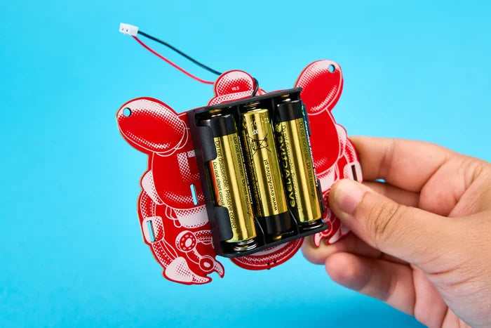 Hand holding a red crab-shaped circuit board with three AA batteries in a black holder against a blue background.