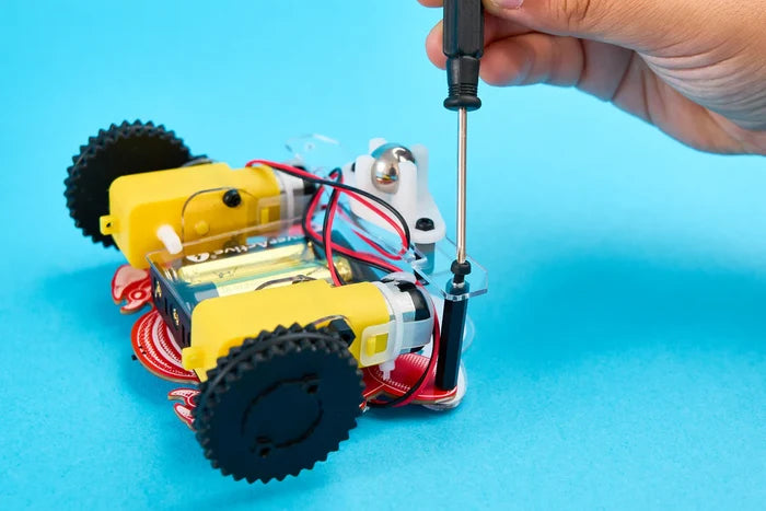 Hand assembling a small robot with yellow motors and black wheels using a screwdriver on a blue background.