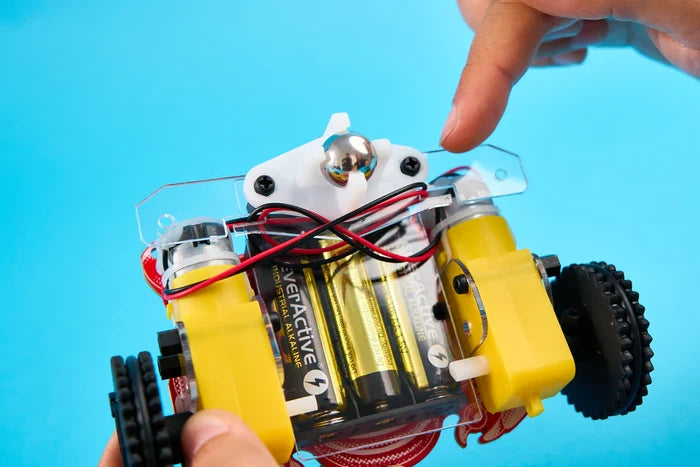 Close-up of a DIY toy robot with yellow motors, black wheels, and a clear frame being held over a blue background.