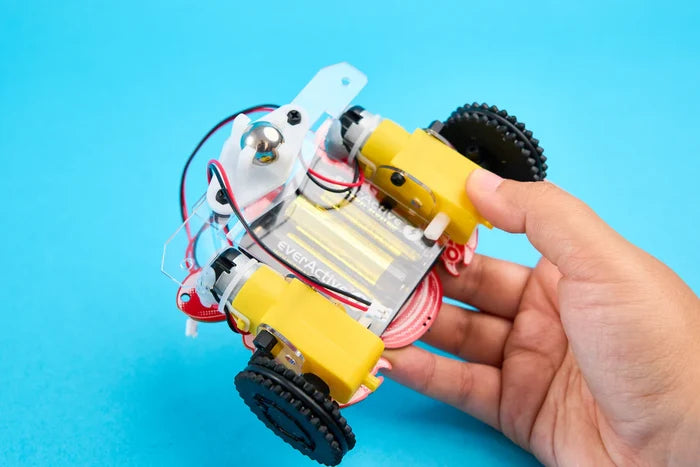 A hand holds a DIY robot with yellow motors, black wheels, clear casing, and red and black wires against a blue background.