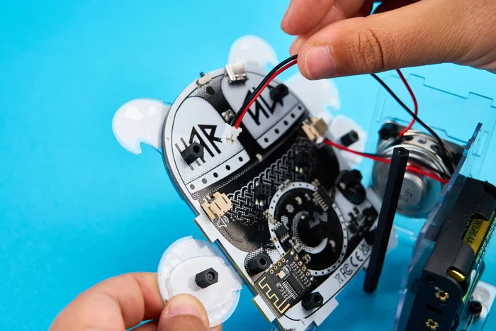 Hands assembling a white, robot-shaped DIY speaker with exposed circuit boards and wires on a bright blue background.