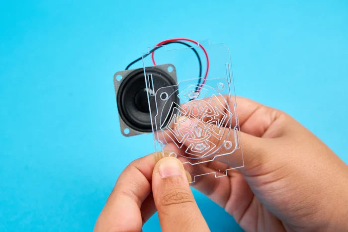Hands hold a clear etched acrylic panel over a small black speaker component against a bright blue background.