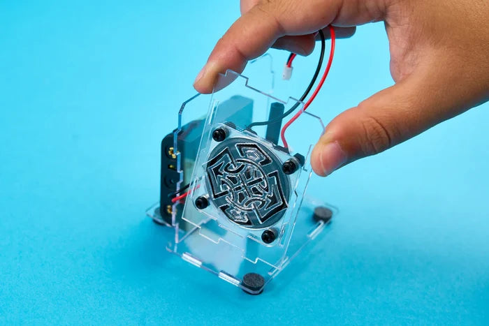 Hand holding a transparent acrylic piece with a decorative metal speaker driver and wires against a blue background.