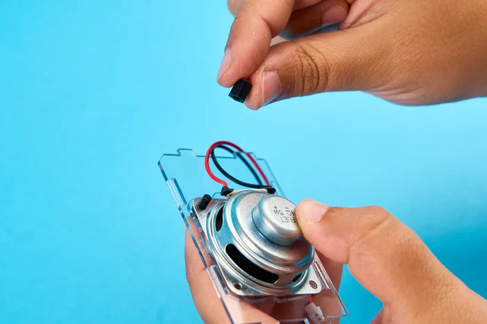 Hands assembling a DIY speaker kit with a metal driver, red and black wires, and a clear plastic mounting board.