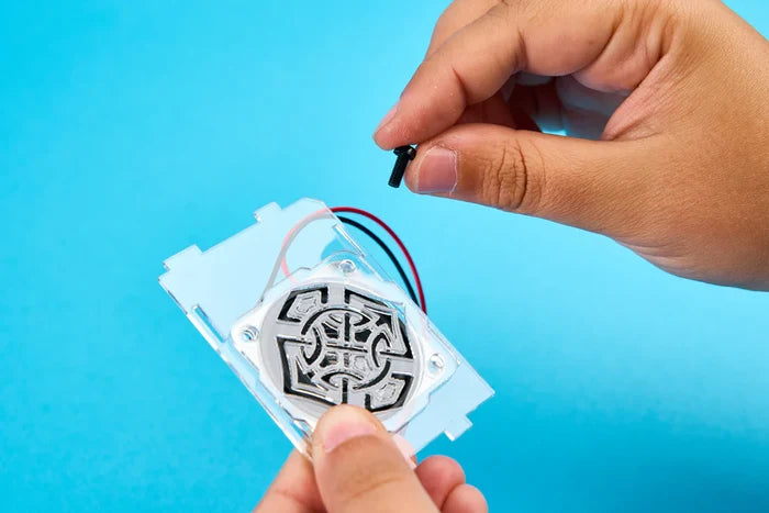 Hands hold a black screw over a small speaker part encased in clear acrylic with red and black wires on a blue background.