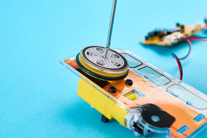 Close-up of a screwdriver assembling a yellow DIY robot toy wheel against a bright blue background.