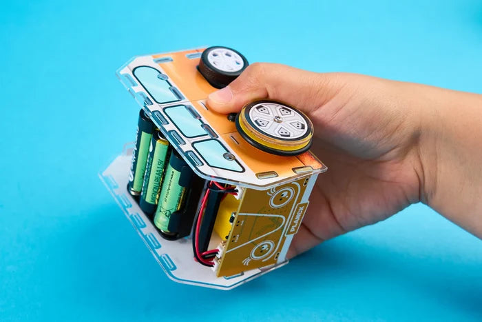 Hand-held yellow and white DIY robot car with visible AA batteries and internal circuitry on a bright blue background.