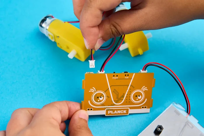 Hands connecting a wire to an orange Planck circuit board with eyes next to yellow motors on a blue background.