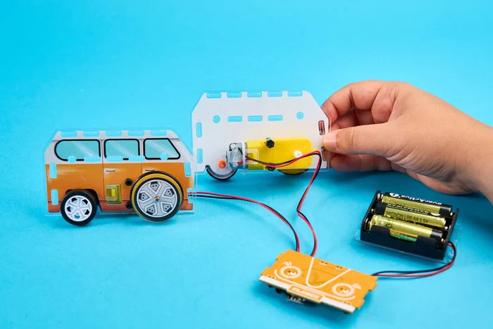 Child's hand assembling an orange toy van with a yellow motor, battery pack, and wires on a blue background.
