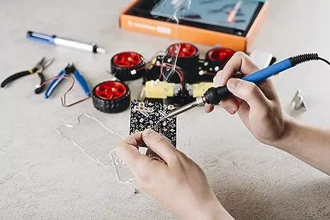 Person soldering a circuit board next to red robot car wheels, tools, and an orange kit box.