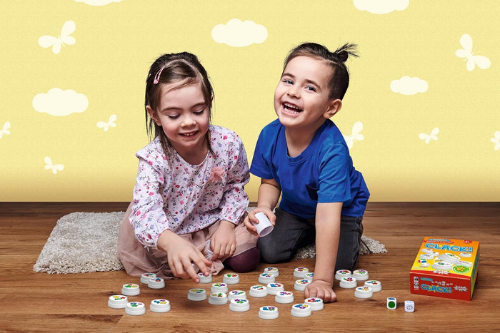 Two children play with white magnetic discs and dice on a wooden floor next to a colorful game box.