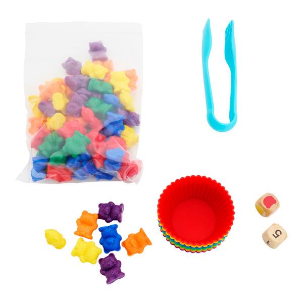 Colorful bear figurines in a bag, blue tweezers, a stack of rainbow silicone cups, and two wooden dice.