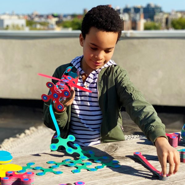 Boy builds a red airplane and green structures using flexible magnetic pieces on an outdoor wooden table.