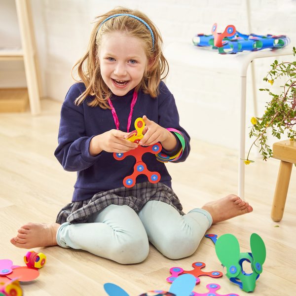 Young girl playing with colorful, flexible magnetic building pieces to create 3D shapes on a wooden floor.