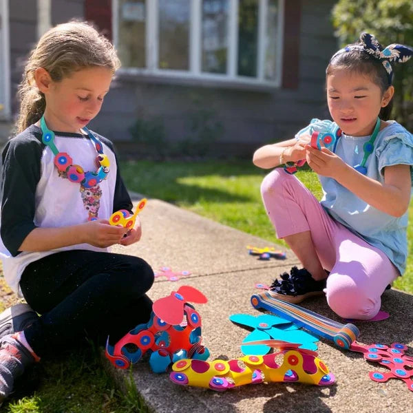 Two girls play outdoors building a toy dog, snake, and necklaces using colorful, flexible magnetic pieces.