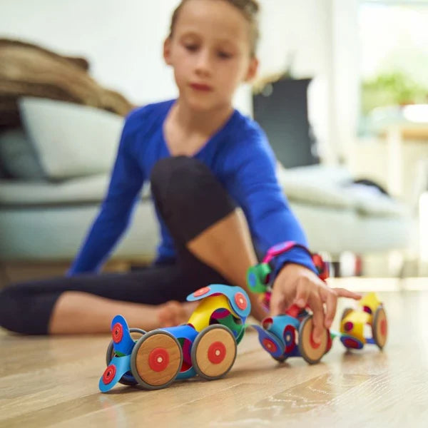 Child building colorful toy vehicles using flexible magnetic strips and wooden wheels on a light floor.