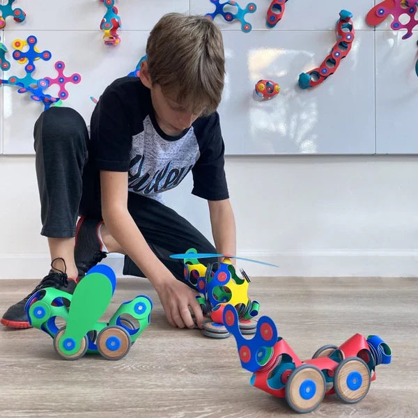 A child builds colorful, flexible magnetic toy vehicles with wheels on a light wood floor.