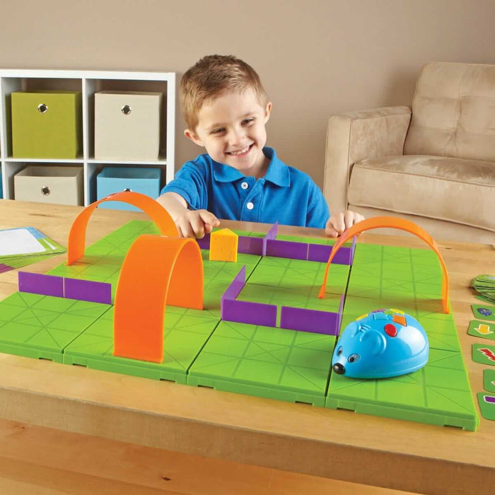 A child plays with a blue robot mouse on a green grid maze with orange tunnels, purple walls, and yellow cheese.