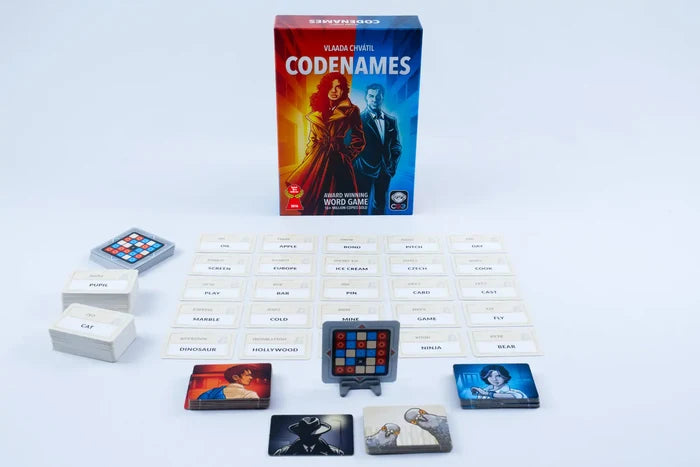 Codenames box with red and blue spy artwork, spread with word cards, colored agent tiles, and grid cards on white.