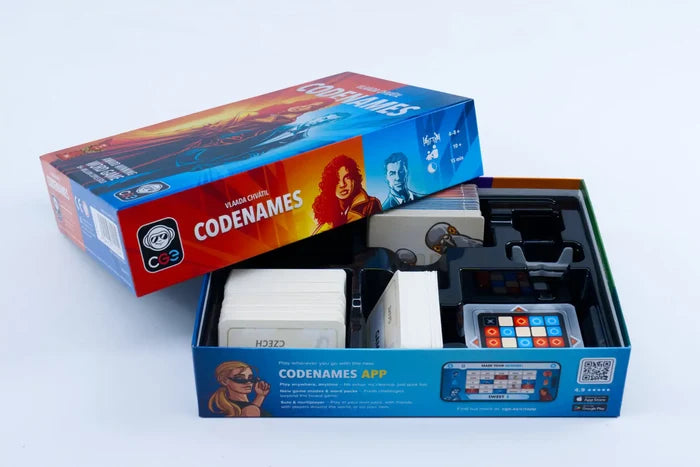 Open red and blue Codenames box showing character art, stacks of white word cards, and game tiles in a black tray.