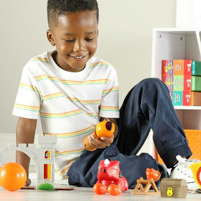 Smiling boy uses an orange wand to play with a red toy dragon, grey castle gate, and treasure chest on the floor.