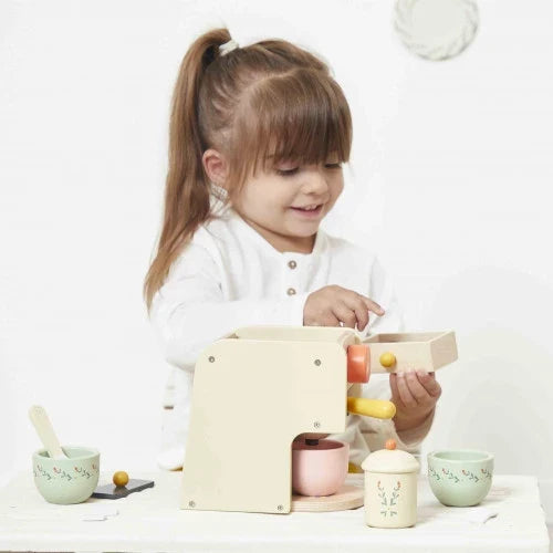 Child playing with a cream-colored wooden toy coffee machine, orange dials, and floral-patterned pastel cups.