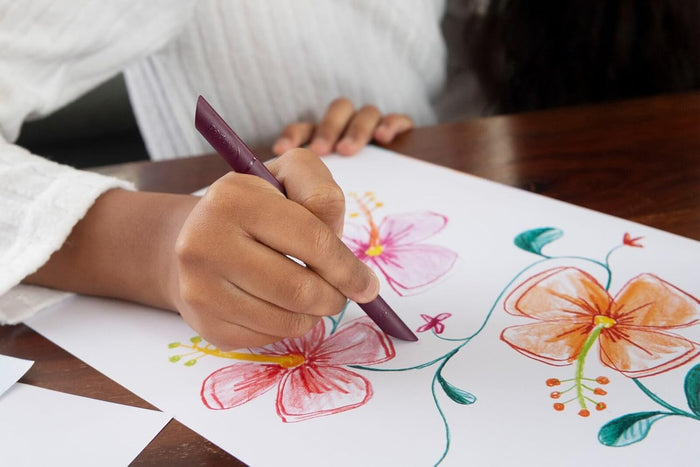 Child's hand using a purple ergonomic crayon to draw vibrant pink and orange flowers on white paper.