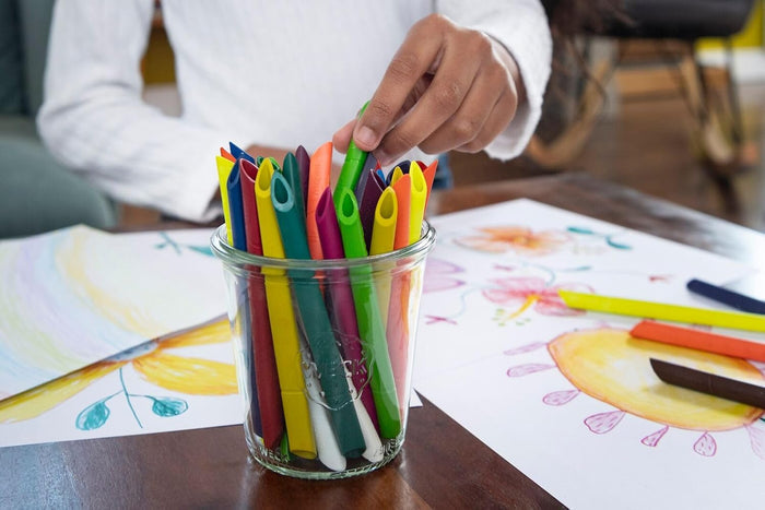 Child reaches for a green triangular crayon from a jar of 24 vibrant crayons on a table with colorful drawings.