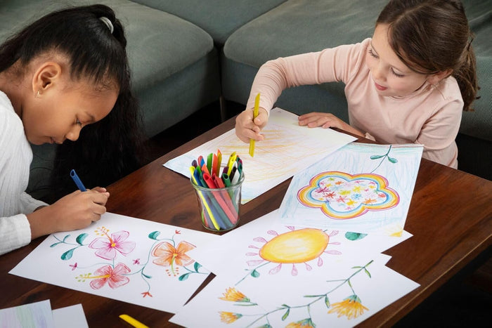Two girls drawing colorful flowers and a sun on paper with bright pencils from a jar on a coffee table.