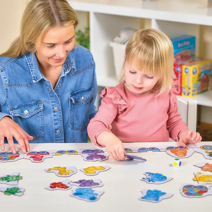 Woman and child match colorful animal pieces to a long train puzzle with a color-coded die on a white table.