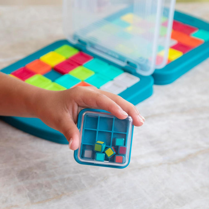 Child's hand holding a small shaker with colored cubes over a teal board with a grid of colorful square tiles.
