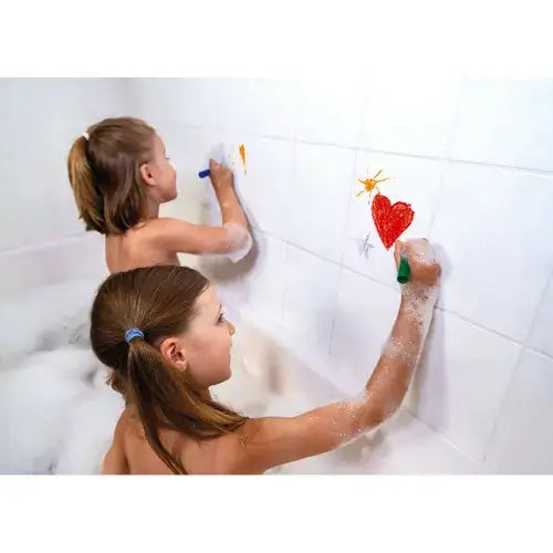 Two girls in a bubble bath drawing a red heart and orange sun on white wall tiles with colorful bath crayons.