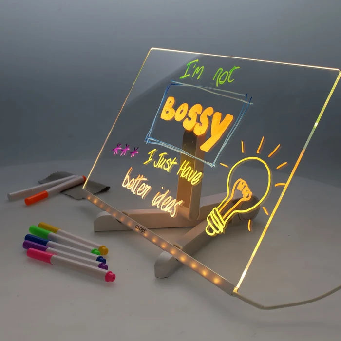 Clear acrylic LED board on a stand with glowing neon writing and drawings, surrounded by colorful markers.