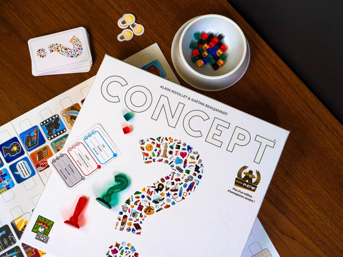 White Concept board game box with a large icon-filled question mark, colorful cubes, and pawns on a wooden table.