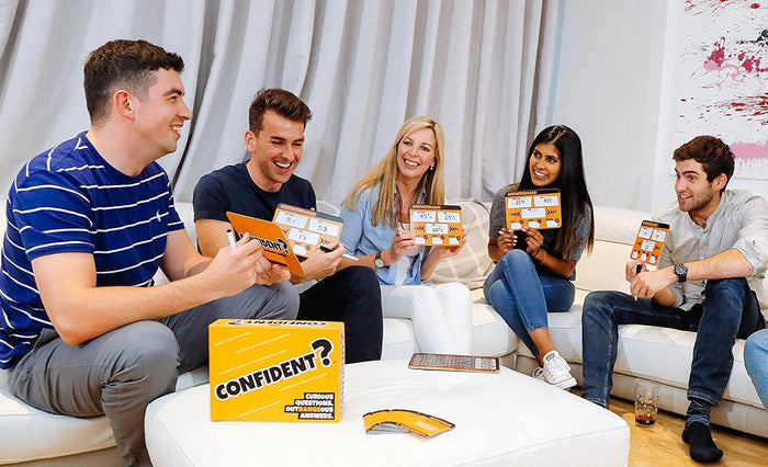 Five adults laugh on a couch playing the CONFIDENT? quiz game with orange answer boards and a bright yellow box.
