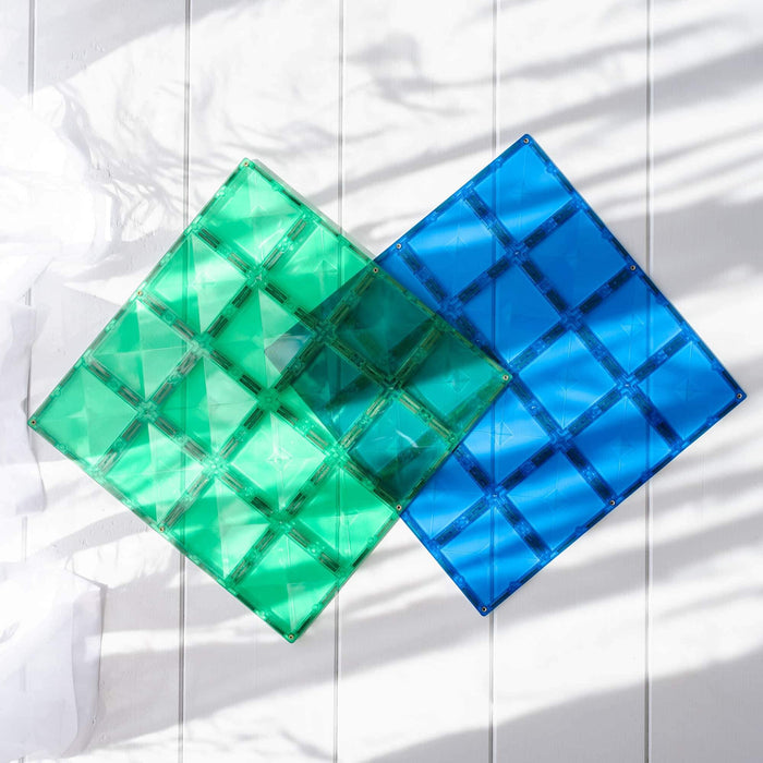 Green and blue square magnetic base plates overlapping on a white wooden surface with soft sunlight and shadows.