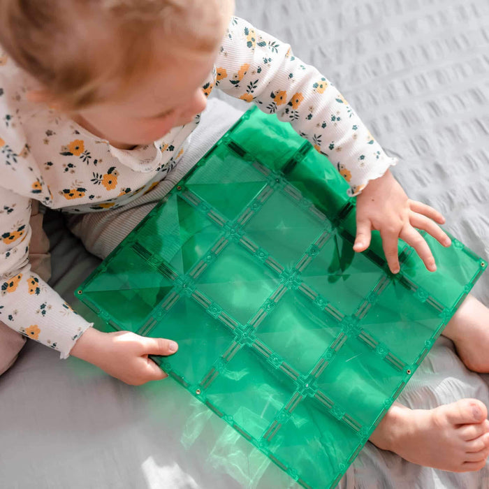 Toddler playing with a large, square, transparent green magnetic tile base plate on a grey textured surface.
