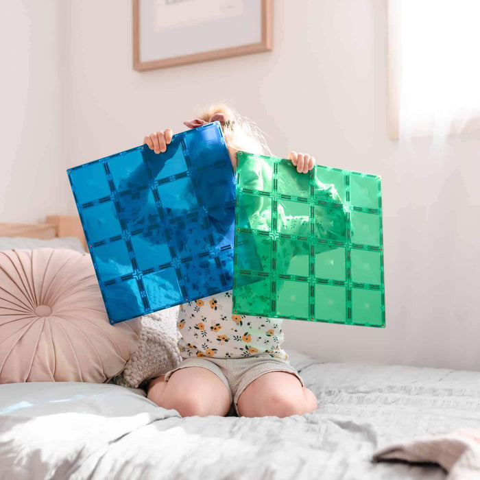 Child holding large blue and green translucent square magnetic base plates in front of their face.
