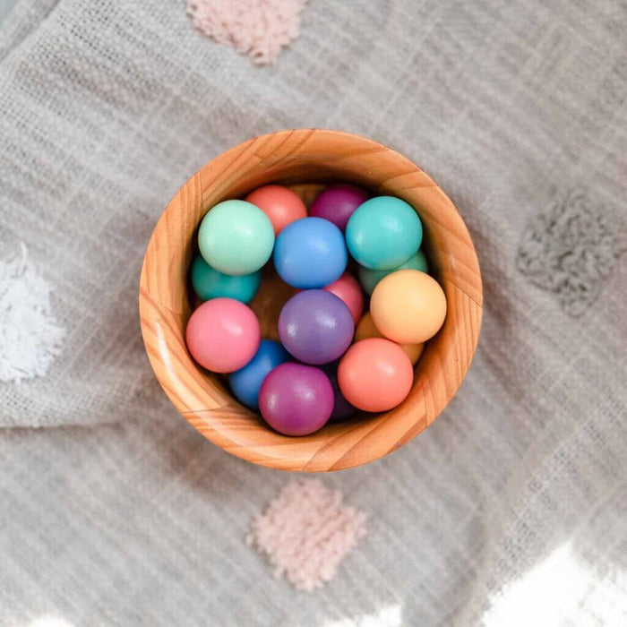 Overhead view of a wooden bowl filled with various pastel-colored wooden balls on a textured grey cloth background.