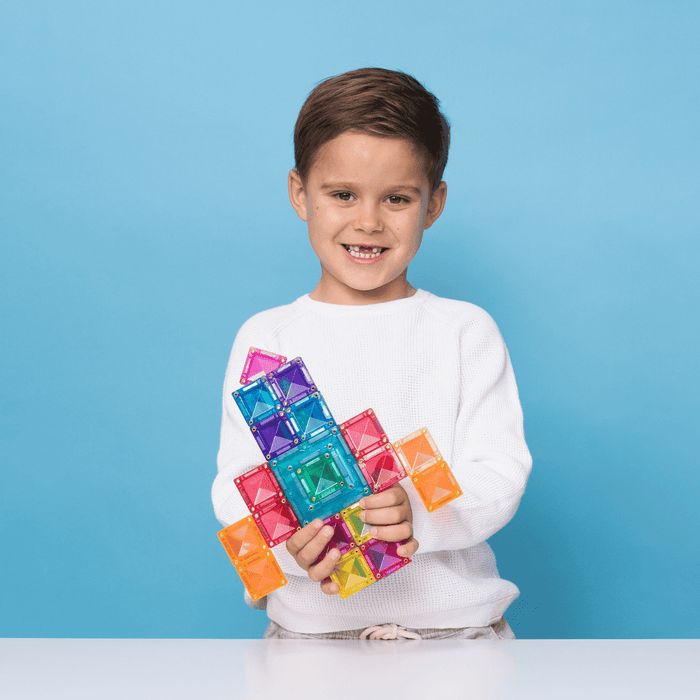 Smiling boy holding a colorful rocket built from bright translucent square and triangle magnetic tiles.