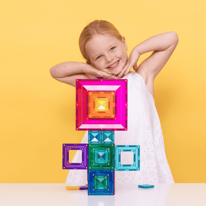 Child smiling behind a flower tower of colorful, translucent magnetic tile frames on a bright yellow background.