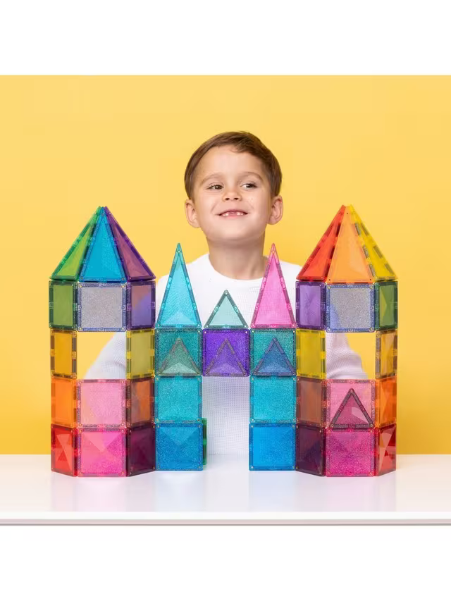 Smiling boy behind a colorful castle built with glittery, translucent magnetic squares and triangles on yellow.