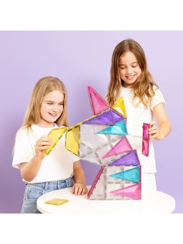 Two girls build a unicorn head using glittery pink, purple, and blue magnetic tiles on a white table.