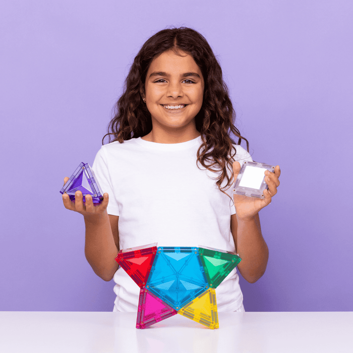 Smiling girl holds magnetic tiles next to a colorful star construction made of translucent geometric shapes.