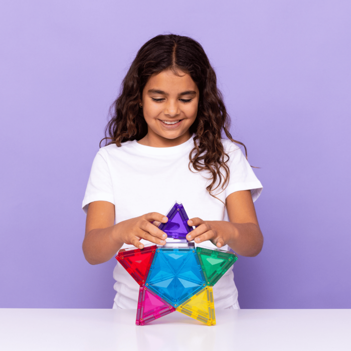 A smiling girl builds a colorful star using translucent magnetic geometric tiles against a light purple backdrop.